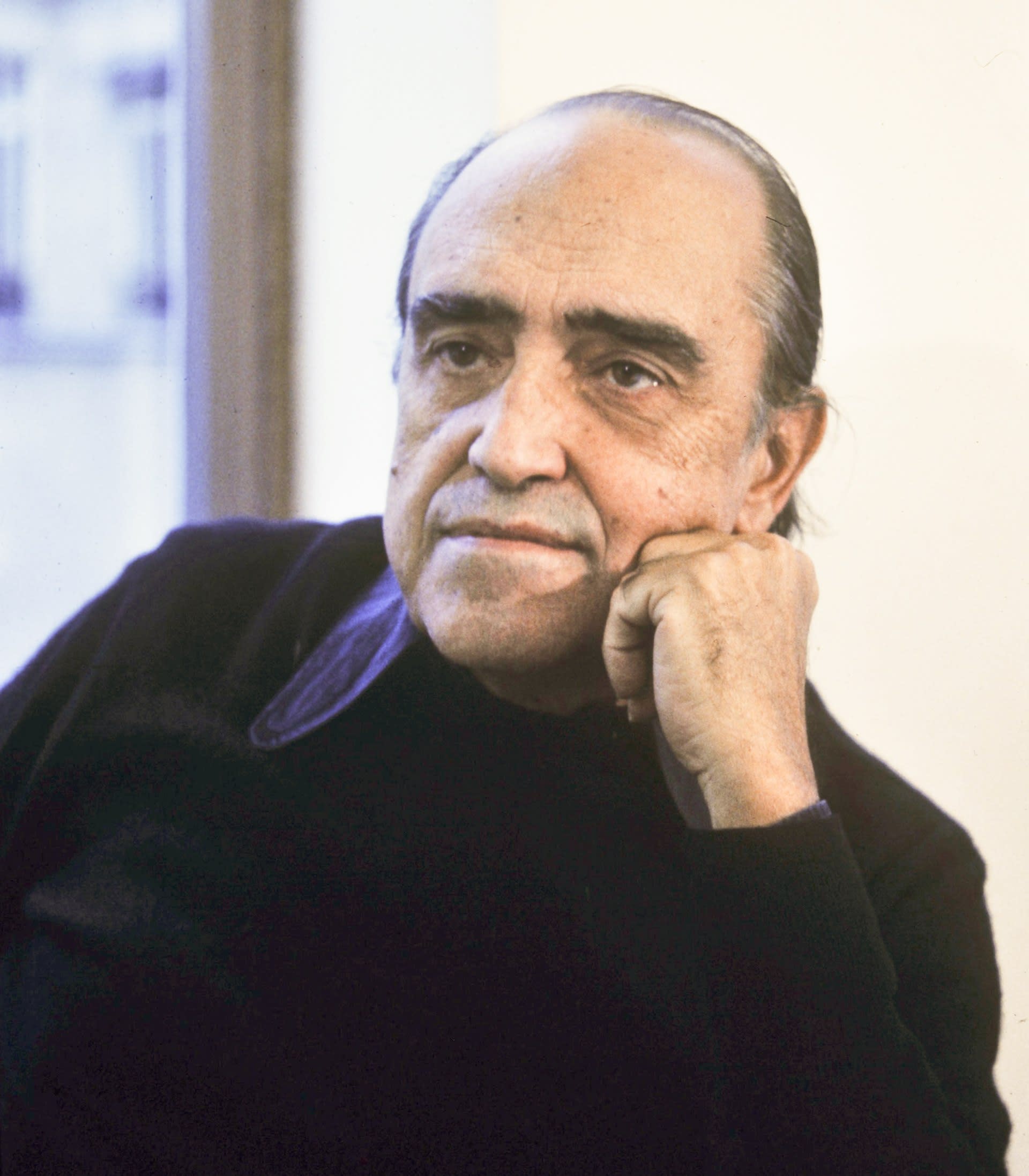 Arquiteto modernista brasileiro Oscar Niemeyer sentado em frente a um modelo de sua obra, com uma vista da cidade ao fundo.